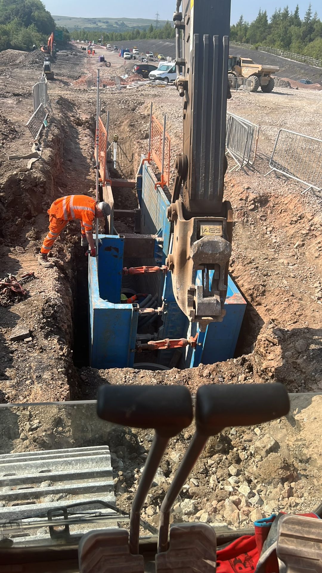 Worker in high-visibility clothing excavating a deep trench with heavy machinery assistance.