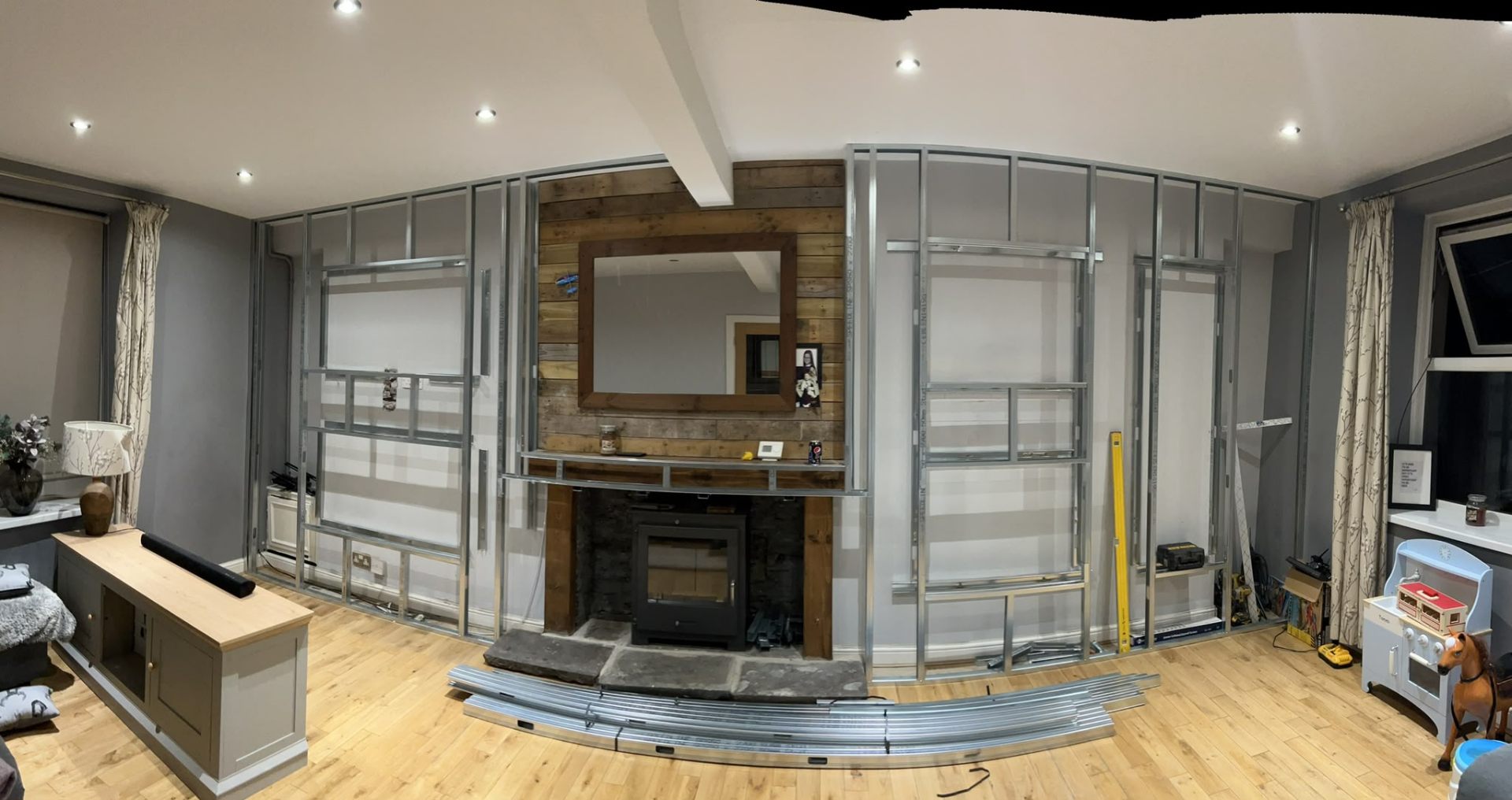 Wide panoramic view of a custom media wall being constructed using metal stud framing around a fireplace.