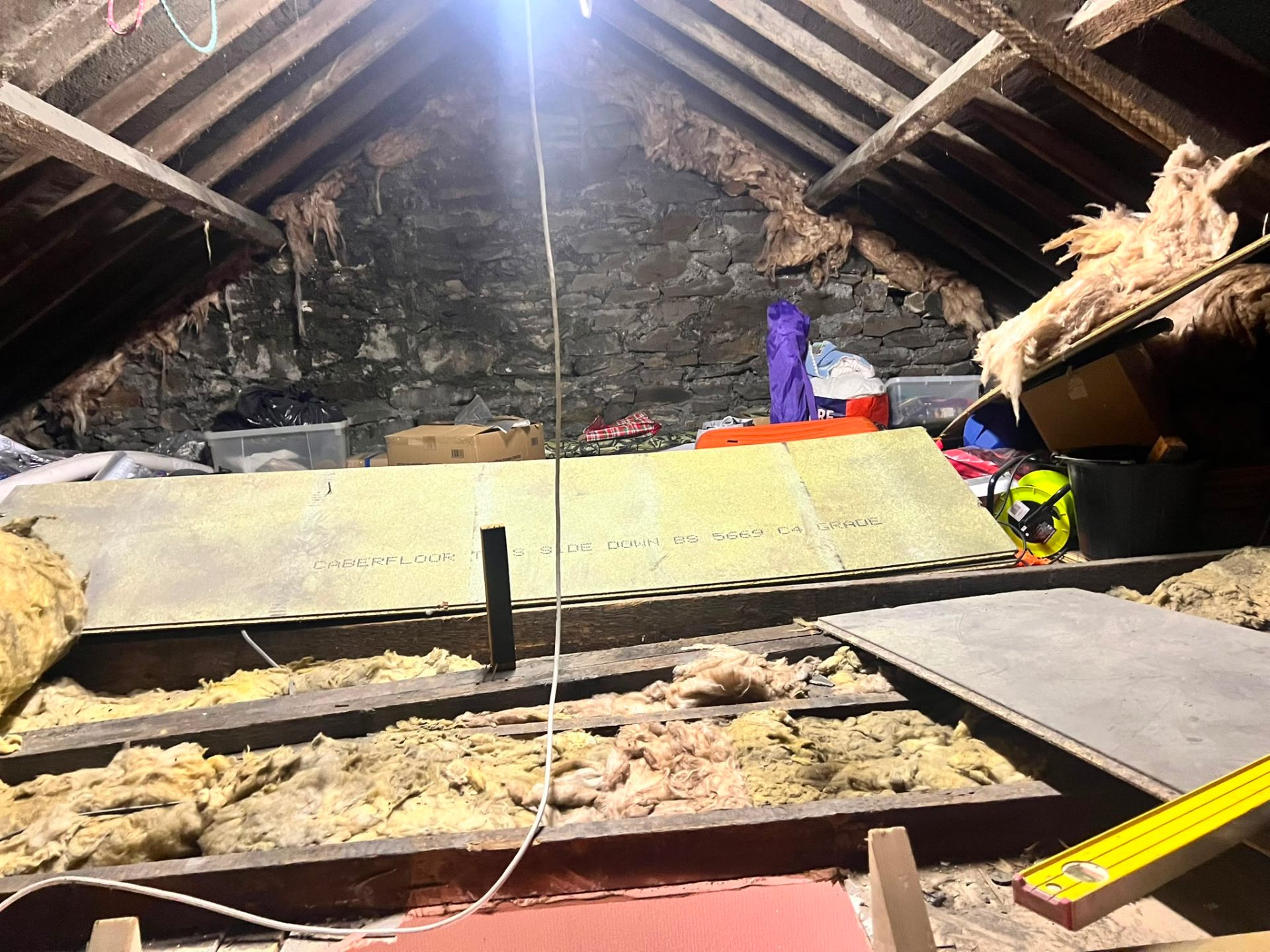 Attic space mid-renovation showing exposed roof rafters, stone walls, and floor insulation materials.