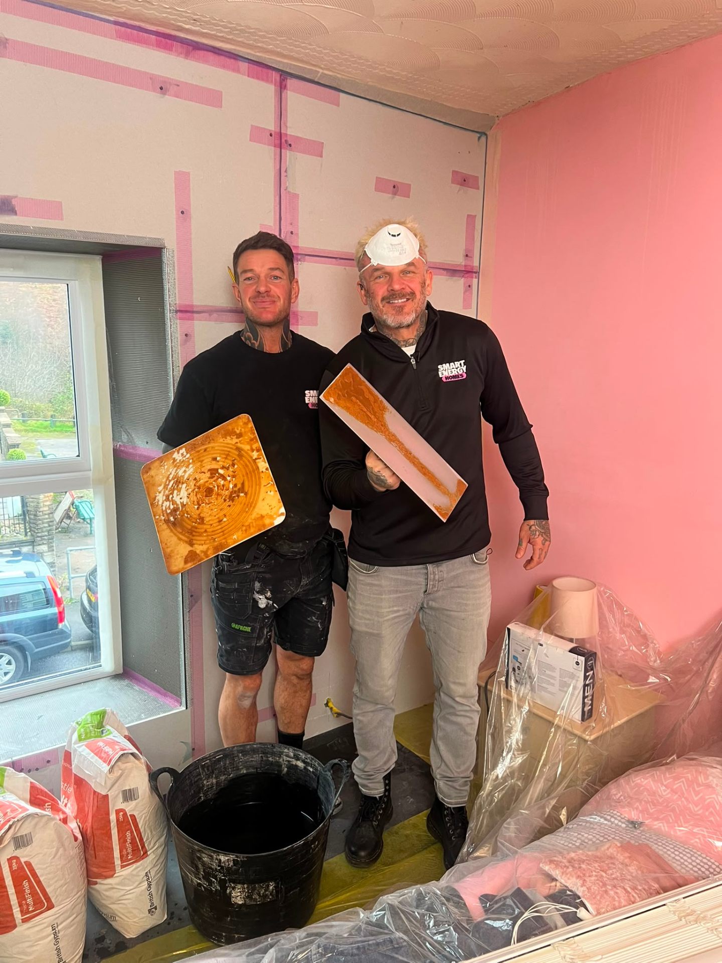 Two plasterers posing with their tools in a room prepared with pink bonding and plasterboard.