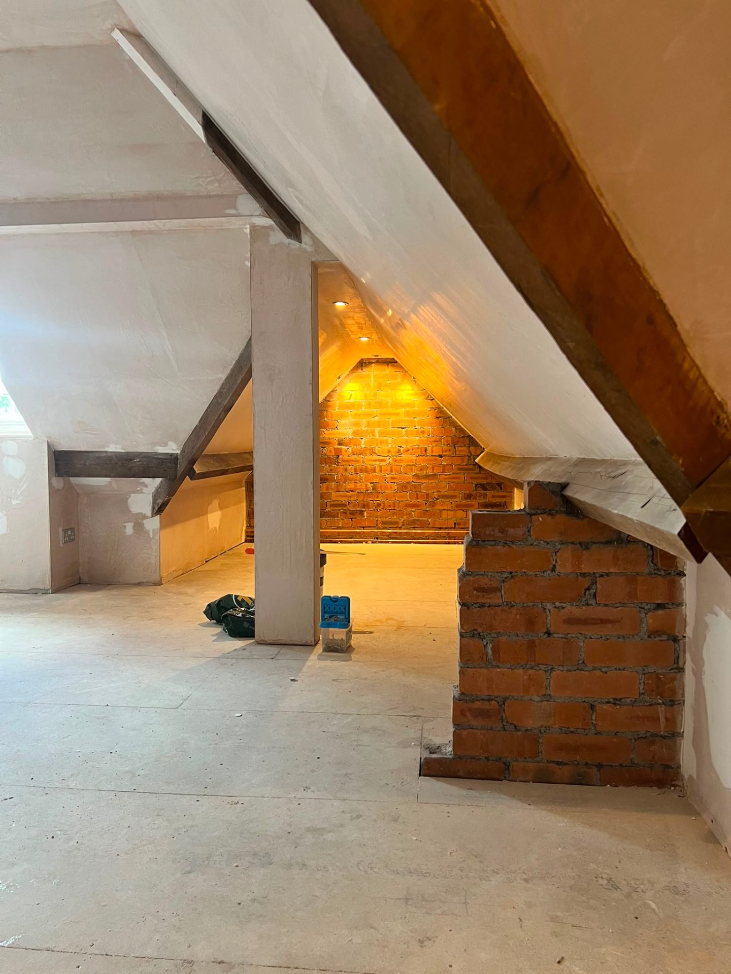 Attic conversion showing structural beams, plastered sloped ceilings, and warm lighting on a brick wall.