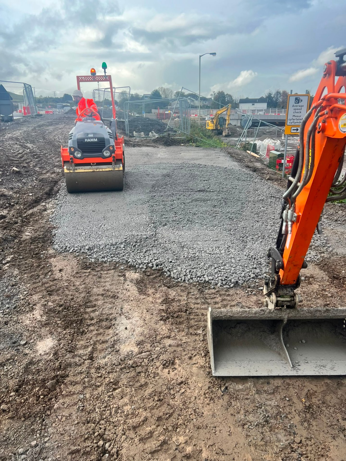 Construction site machinery including a road roller and excavator bucket leveling a gravel surface.